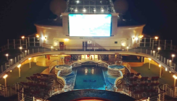 A luxury cruise ship deck featuring a pool surrounded by lounge chairs and warm ambient lighting. A large screen overlooks the area, showing an image against a dark night sky.