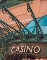 The image displays the entrance to a casino, characterized by detailed and ornate architectural elements with green metal structures and a sign that reads 'CASINO'. The background includes a glass dome and a peach-colored wall.