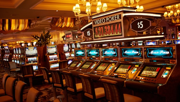 A casino floor featuring rows of brightly lit video poker machines. The area is illuminated by elegant chandeliers, with a display board showing a large sum of money, suggesting a possible jackpot. The rich decor includes luxurious carpets and comfortable seating, adding to the opulent atmosphere.