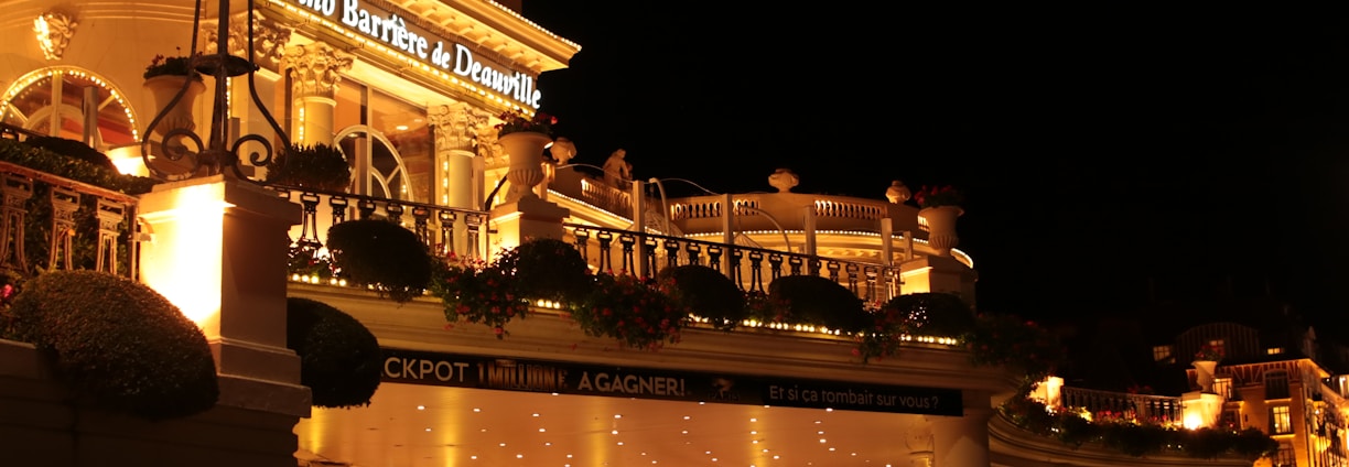 A luxurious casino at night, illuminated with warm, golden lights. The building features elegant architecture with columns and decorative elements. A few people are walking towards the entrance, and there are a couple of parked cars nearby.