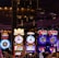 A lively casino scene featuring a row of bright, colorful slot machines with themes like Monopoly, Michael Jackson, and Lord of the Rings. The gaming machines display a variety of flashing lights and digital screens, creating an energetic and exciting atmosphere. Reflections of the lights are visible in glossy surfaces above the machines.