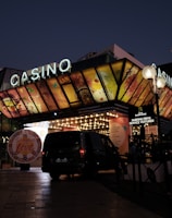 A building with a brightly illuminated entrance is seen at night. The sign above reads 'Casino' in bright letters. The facade features colorful graphics depicting various indulgent themes. Several vehicles are parked near the entrance, and people are visible in the vicinity. Tall lamp posts with spherical lights are present, casting a warm glow.