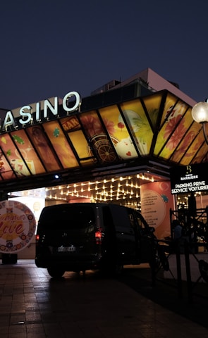A building with a brightly illuminated entrance is seen at night. The sign above reads 'Casino' in bright letters. The facade features colorful graphics depicting various indulgent themes. Several vehicles are parked near the entrance, and people are visible in the vicinity. Tall lamp posts with spherical lights are present, casting a warm glow.