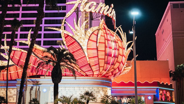 A brightly lit casino with neon lights in vibrant red, pink, and blue, featuring a large flamingo signage. Palm trees are visible in the foreground with vehicles passing by on the street at night.