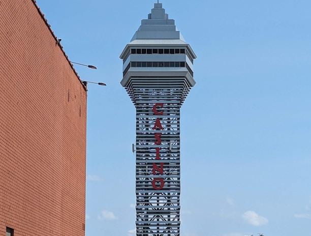 A tall tower with a modern design features the large vertical red letters spelling 'CASINO'. The structure has a sleek, metallic appearance with various horizontal and vertical lines. The sky is mostly clear with a few clouds. In the foreground, part of a brick building is visible, along with some trees and buildings in the distance.